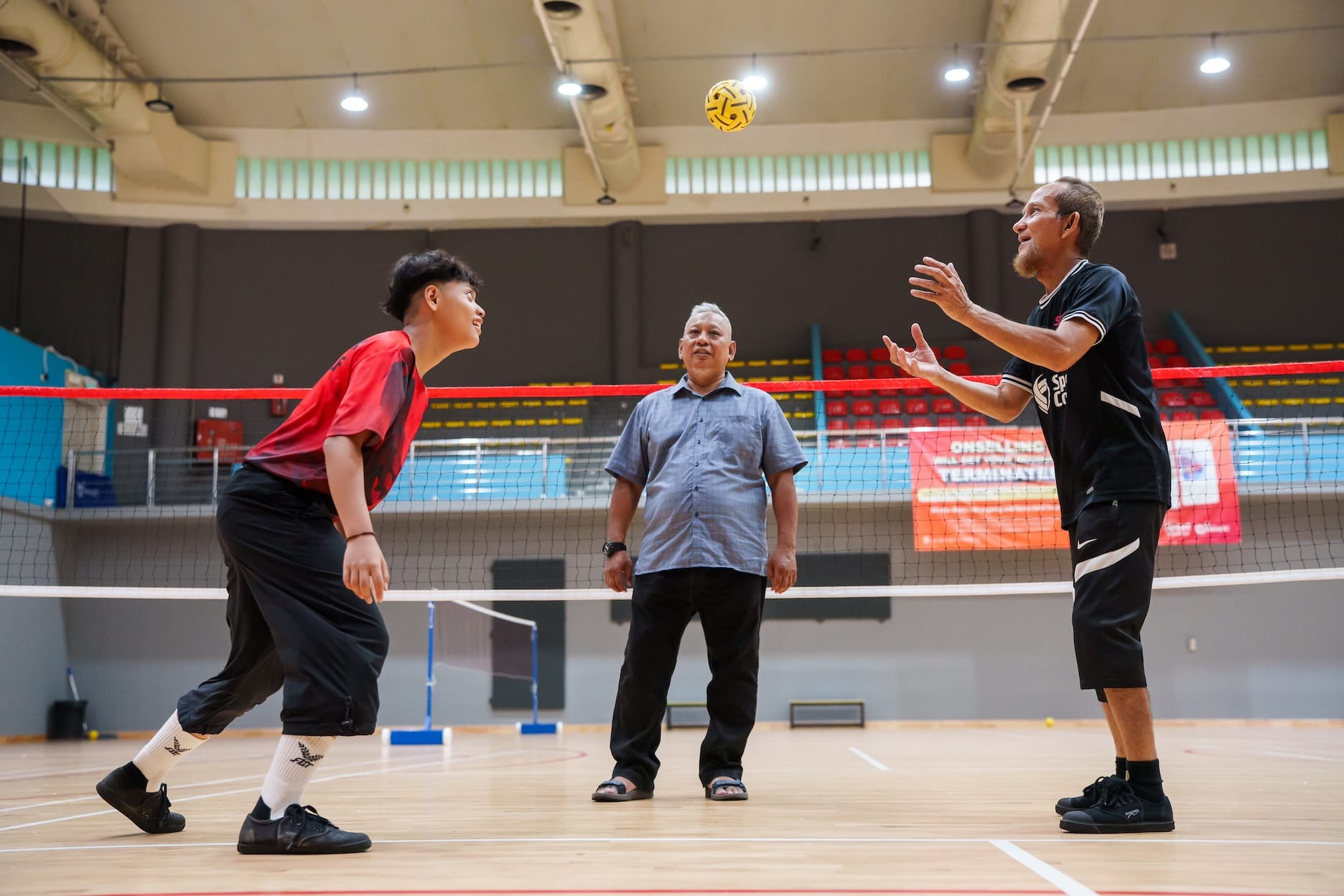 Three men playing sepak takraw; yellow ball in mid-air above net.